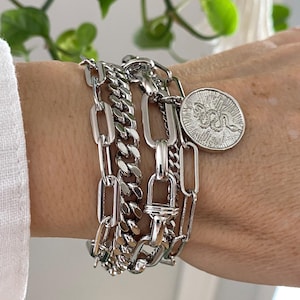 May include: A collection of silver-toned bracelets with various chain link designs. One bracelet features a coin charm with a snake emblem. The bracelets are stacked together, creating a layered look. The image is well-lit, highlighting the metallic shine.