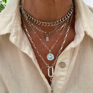 May include: A layered silver-toned necklace set featuring a thick curb chain, a rectangular link chain with a small blue pendant, a delicate beaded chain with a circular turtle charm in light blue, and a long chain with a large oval link pendant.