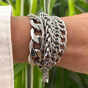 May include: A multi-layered silver-toned metal chain bracelet featuring a combination of thick, polished curb links and finer, intricate woven chain strands. The bracelet has a high-shine metallic finish and is secured with a small metal clasp.