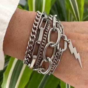 May include: A multi-strand silver-toned bracelet featuring a combination of various chain styles, including thick oval links, flat curb chains, and a prominent lightning bolt charm dangling from the side, all crafted from polished metallic material.
