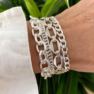 May include: A stack of three silver-toned chain bracelets worn on a wrist. The set includes a chunky oval link chain, a smaller textured curb chain, and a central chain featuring elongated rectangular links with a polished metallic finish.