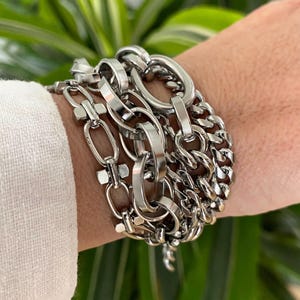 May include: Several silver-coloured bracelets with different chain link styles. The bracelets feature oval links, curb chains, and other geometric forms. The jewellery is stacked on a wrist, creating a layered effect. Green foliage is in the background.