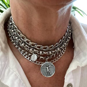 Peut inclure: Un collier argenté à plusieurs rangs avec différents styles de chaînes. Le collier comprend un pendentif pièce et un pendentif rond avec un dessin de figure humaine. Le bijou est porté sur une chemise blanche.
