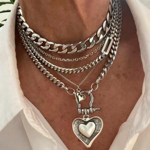 Peut inclure: Ensemble de colliers argentés superposés. Les colliers présentent différents styles, notamment des chaînes à maillons épais, des chaînes délicates et un pendentif en forme de cœur. Les bijoux sont portés sur une chemise blanche.