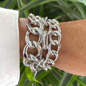 May include: A silver chain bracelet featuring large, interlocking links. The bracelet is made of a shiny metal and is worn on the wrist. The bracelet is a bold statement piece.