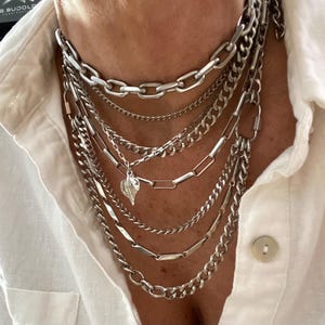 May include: A collection of layered silver necklaces. The necklaces feature various chain styles, including curb, cable, and paperclip chains. A small leaf pendant hangs from one of the chains. The necklaces are worn over a white button-down shirt.