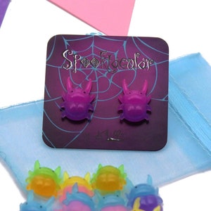 May include: A set of 10 colorful glow-in-the-dark crab earrings. The earrings are packaged on a purple card with a spiderweb design and the text "Spooktacular".