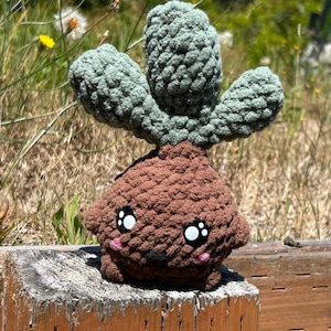 Pode incluir: Um peluche de cacto de crochê feito à mão, com uma base marrom e almofadas de cacto verdes. O peluche tem olhos grandes e bochechas rosadas. Está colocado em um poste de madeira em um ambiente natural.