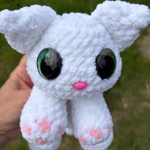May include: A white crocheted plush animal with large, round, black and green glittery eyes. The toy features a small pink plastic nose and light pink stitched paw pads on its front feet. The texture is soft, fuzzy, and made of yarn.