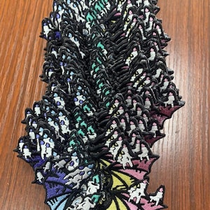 May include: A stack of rainbow dragon embroidered iron-on patches. The patches are in a variety of colors, including pink, yellow, green, blue, and purple. The dragon is facing to the left and has a long tail.
