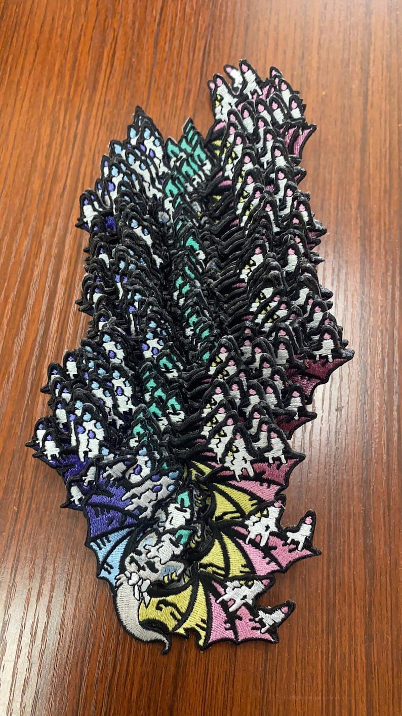 May include: A stack of rainbow dragon embroidered iron-on patches. The patches are in a variety of colors, including pink, yellow, green, blue, and purple. The dragon is facing to the left and has a long tail.