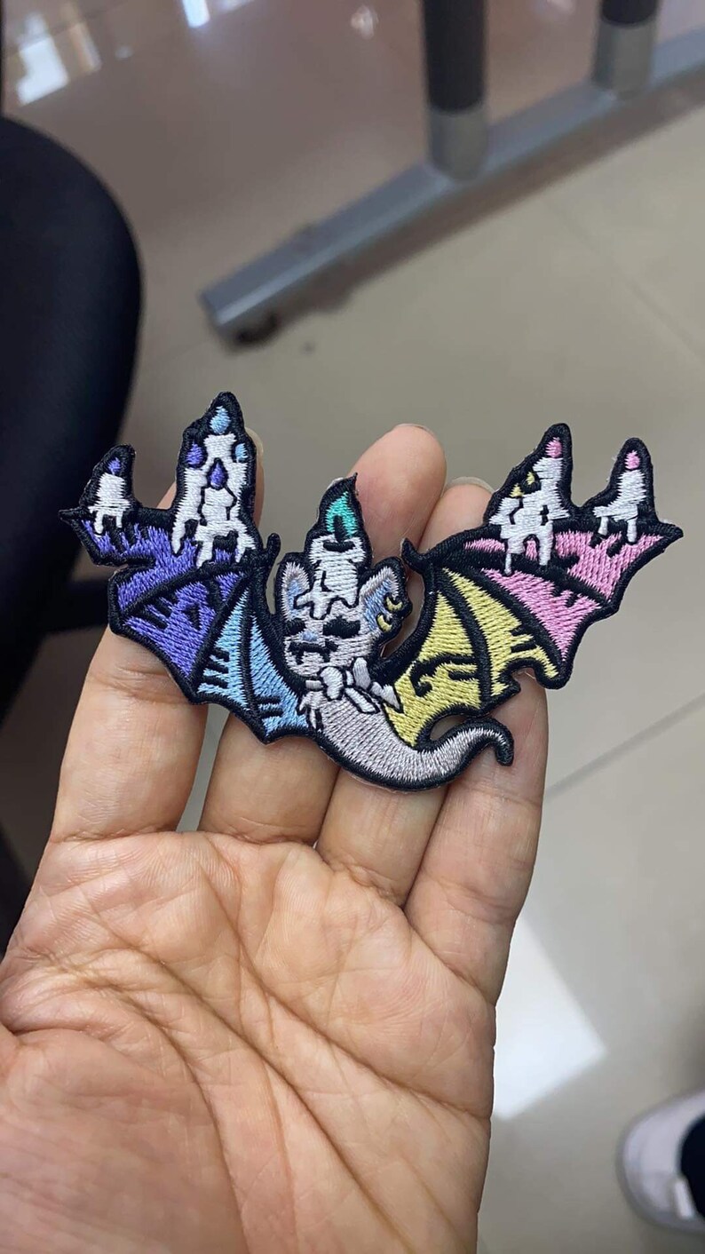 May include: An embroidered iron-on patch featuring a white bat with rainbow wings. The bat is wearing a party hat and has a cute, cartoon-like expression.
