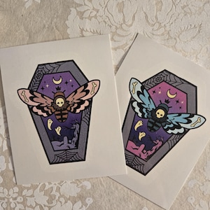 May include: Two sticker designs featuring a moth with a skull head inside a purple coffin with a spiderweb design. The moth on the left is pink and brown, and the moth on the right is blue and green.