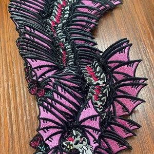 May include: A stack of pink and black embroidered patches featuring a bat with wings spread wide. The bat has a detailed design with a skull-like face and roses.