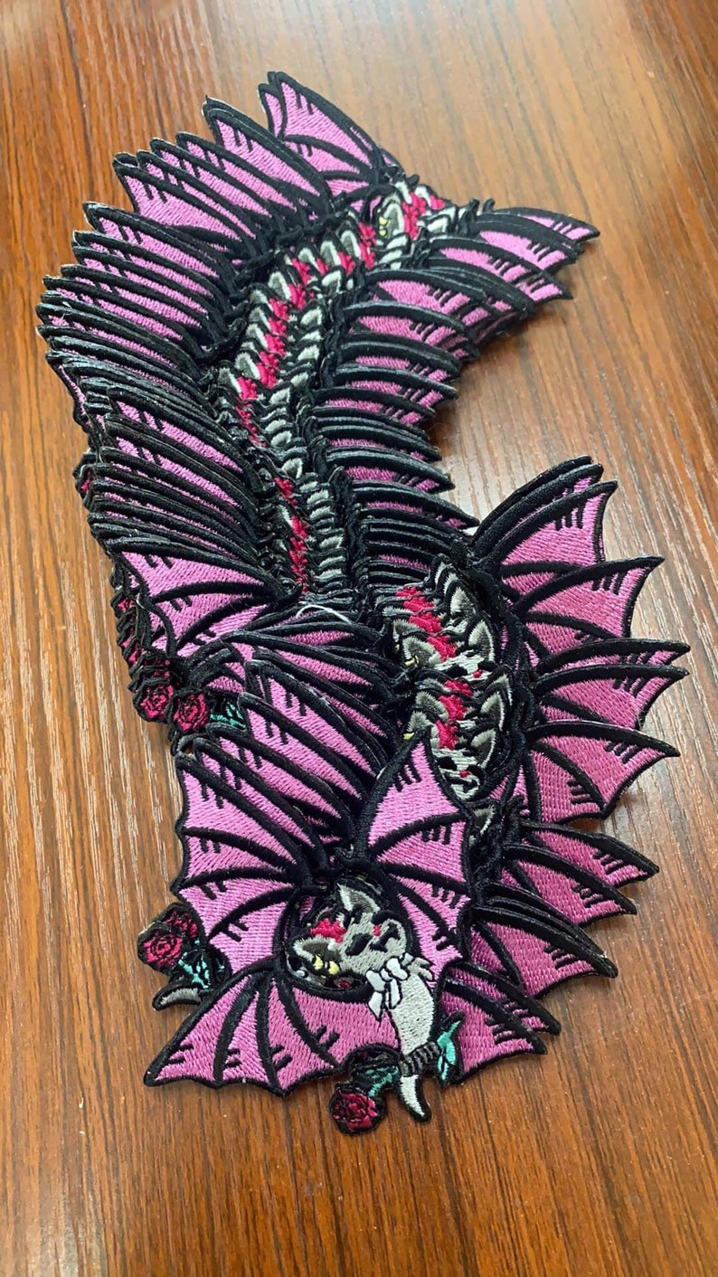 May include: A stack of pink and black embroidered patches featuring a bat with wings spread wide. The bat has a detailed design with a skull-like face and roses.