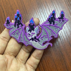 May include: Purple embroidered iron-on patch of a bat with wings spread wide. The bat is holding a black candle with a blue flame. The bat is surrounded by three black candles with blue flames.