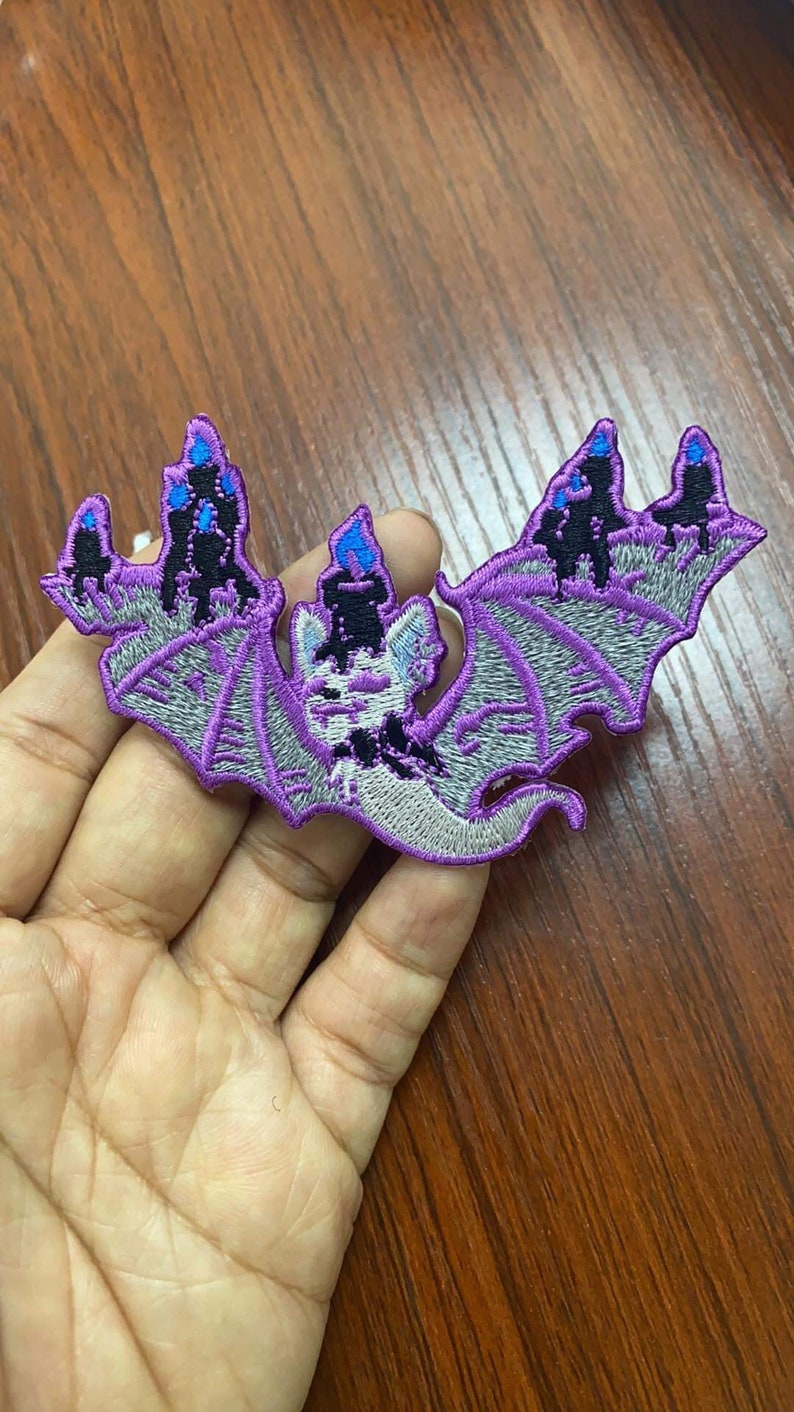 May include: Purple embroidered iron-on patch of a bat with wings spread wide. The bat is holding a black candle with a blue flame. The bat is surrounded by three black candles with blue flames.
