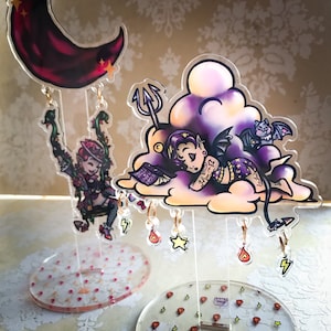 May include: Two acrylic charms featuring a purple and black demon character sleeping on a cloud and a purple and green character with wings. Both charms are attached to clear acrylic stands with dangling charms.