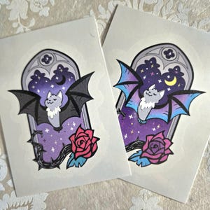 May include: Two stickers featuring a cute cartoon bat with a pink or blue gradient, flying in front of a purple starry night sky with a crescent moon. The bat is inside a gothic archway with a thorny rose vine.