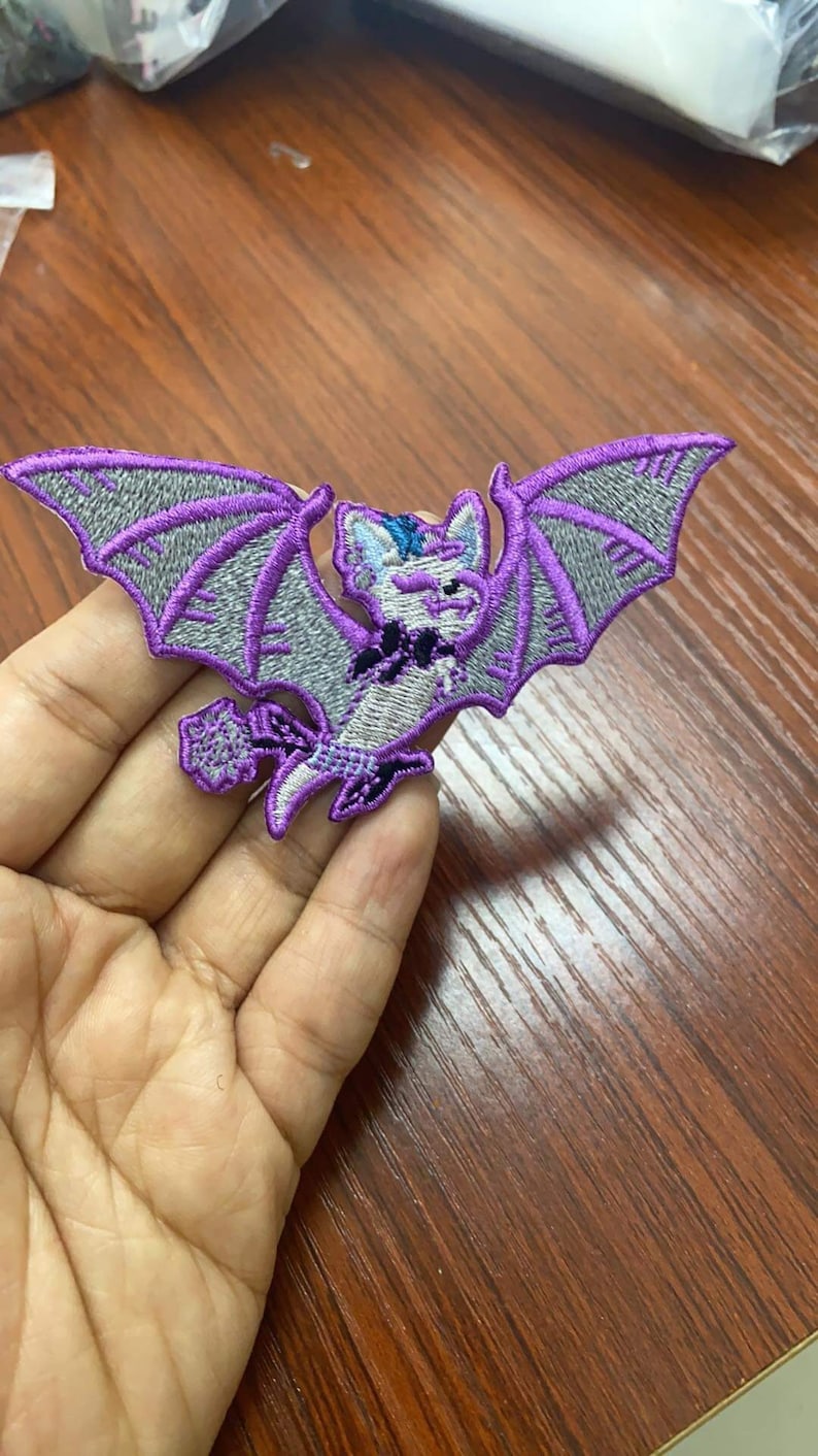 May include: Purple and gray embroidered iron-on patch of a cartoon bat with a black and white striped scarf.