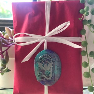 May include: A red gift box tied with white ribbon and a decorative wax seal featuring a bat and a gothic archway design. The seal has a teal, purple, and blue colour scheme.