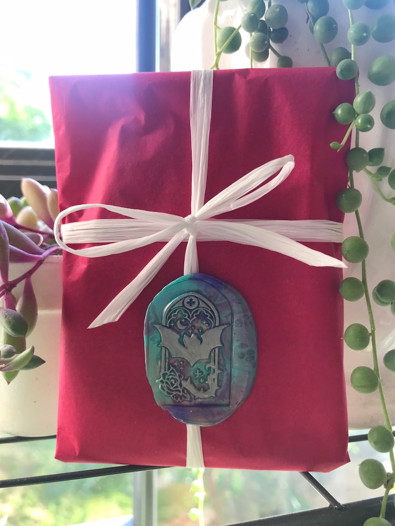 May include: A red gift box tied with white ribbon and a decorative wax seal featuring a bat and a gothic archway design. The seal has a teal, purple, and blue colour scheme.