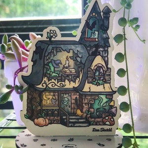 May include: A hand-painted wooden cutout of a whimsical cottage with a witch's hat roof, a glowing window, and a cauldron. The cottage is on a round wooden base with a repeating pattern of stars and moons. The base is labelled "Zina Sketch!"