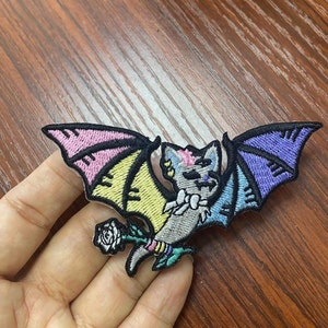 May include: A colorful embroidered iron-on patch featuring a cartoon bat with rainbow wings and a white rose. The bat is gray with pink, yellow, blue, and purple accents.