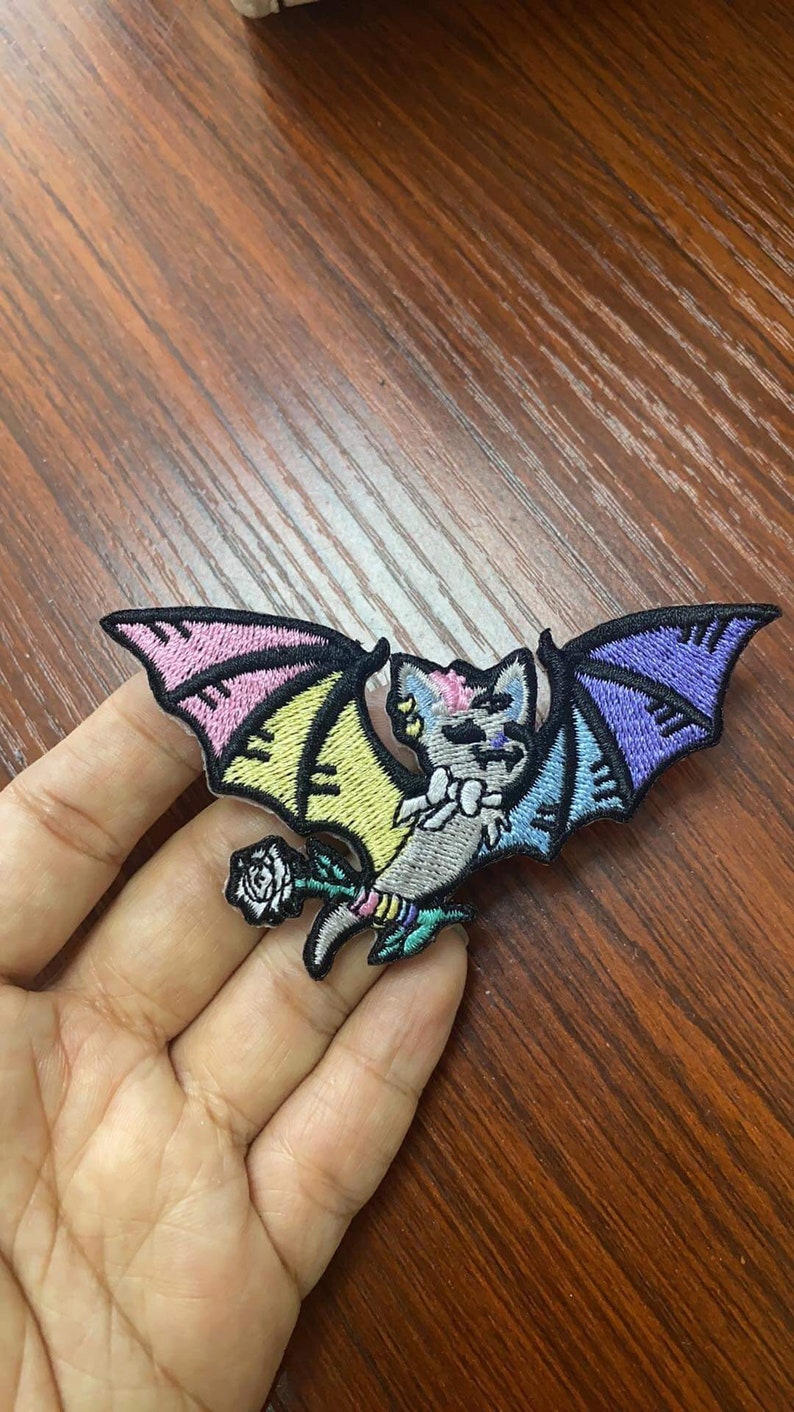 May include: A colorful embroidered iron-on patch featuring a cartoon bat with rainbow wings and a white rose. The bat is gray with pink, yellow, blue, and purple accents.