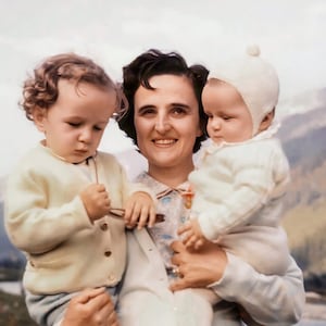 Może przedstawiać: Zdjęcie w stylu vintage przedstawiające kobietę trzymającą dwoje małych dzieci. Kobieta ma na sobie sukienkę w kwiaty i jasny kardigan. Dzieci ubrane są w swetry i czapki. W tle widać rzekę i góry.