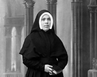 May include: A full-length, black and white photograph of a person in a traditional religious habit. The habit includes a long, pleated skirt, a hooded cloak, and a white wimple framing the face. The person holds a small book.