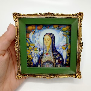 May include: A framed painting of a woman with a blue and white robe, surrounded by flowers and fruit. The frame is gold with a green mat.