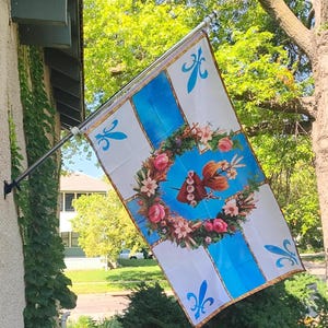 May include: A rectangular flag with a white background and blue cross design, adorned with fleur-de-lis symbols. The center features a floral wreath with a butterfly and a heart-shaped design. The flag is mounted on a pole.