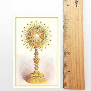 May include: A rectangular print featuring a detailed illustration of a gold monstrance with a central white circular element, surrounded by radiating rays and colorful accents. A wooden ruler is placed alongside, showing a height of 5.5 inches.