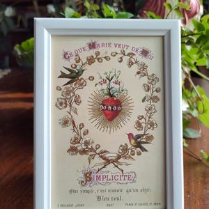 May include: A framed print with a floral wreath surrounding a red heart with a golden halo. The text in the image reads "Ce que Marie veut de toi" and "Simplicité. Etre simple, c'est n'avoir qu'un objet: Dieu seul."  The bottom of the print reads "E Bouasse Jeune 3367 Place St Sulpice, 12, Paris."