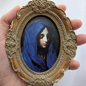 May include: A framed portrait of a woman wearing a blue shawl. The frame is oval shaped and has a gold finish with ornate details.