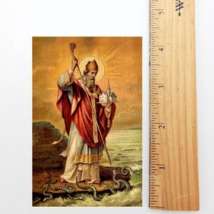 May include: Vintage religious print featuring a saint in red and white robes, holding a staff and a miniature church. The saint stands on a rock with green snakes at his feet, set against a golden background. The print is approximately 3 inches tall.