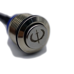Puede incluir: Un interruptor de bot&oacute;n pulsador plateado con un cable negro y rojo conectado. El bot&oacute;n tiene un dise&ntilde;o circular grabado con una espada y una corona de laurel.