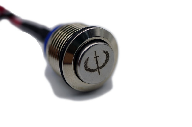 Puede incluir: Un interruptor de bot&oacute;n pulsador plateado con un cable negro y rojo conectado. El bot&oacute;n tiene un dise&ntilde;o circular grabado con una espada y una corona de laurel.