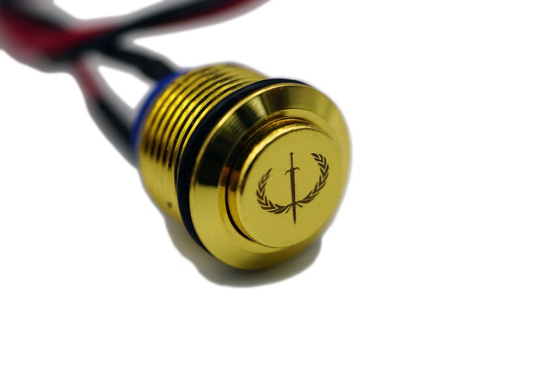 Puede incluir: Un bot&oacute;n pulsador de color dorado con un logotipo en blanco y negro de una espada con una corona de laurel alrededor.