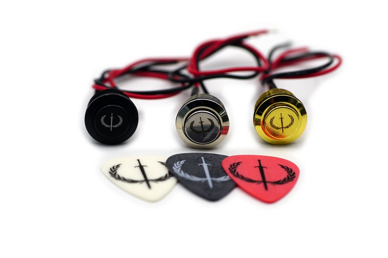 Puede incluir: Tres botones de guitarra con acabado negro, plateado y dorado. Cada bot&oacute;n presenta un logotipo con una espada y una corona de laurel. Los botones se acompa&ntilde;an de tres p&uacute;as de guitarra con el mismo logotipo en blanco, negro y rojo.