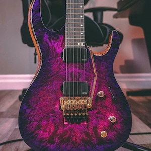 Puede incluir: Una guitarra el&eacute;ctrica p&uacute;rpura y negra con un puente y perillas doradas. La guitarra tiene un patr&oacute;n &uacute;nico de veta de madera en espiral.