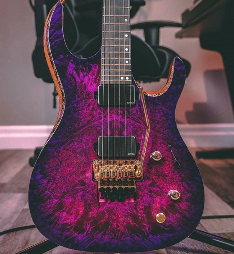 Puede incluir: Una guitarra el&eacute;ctrica p&uacute;rpura y negra con un puente y perillas doradas. La guitarra tiene un patr&oacute;n &uacute;nico de veta de madera en espiral.