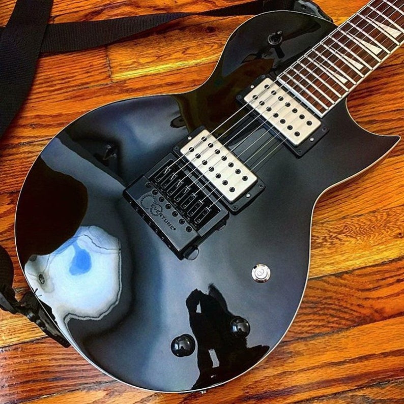 Puede incluir: Una guitarra el&eacute;ctrica negra con una sola pastilla y una correa negra. La guitarra tiene un acabado negro y un diapas&oacute;n de palisandro.