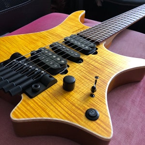 Puede incluir: Una guitarra el&eacute;ctrica amarilla con una golpeador negro y una perilla negra. La guitarra tiene un cuerpo en forma de estrella &uacute;nico y un puente negro.