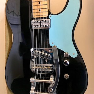 Puede incluir: Guitarra el&eacute;ctrica negra con una golpeador azul claro y un vibrato Bigsby.