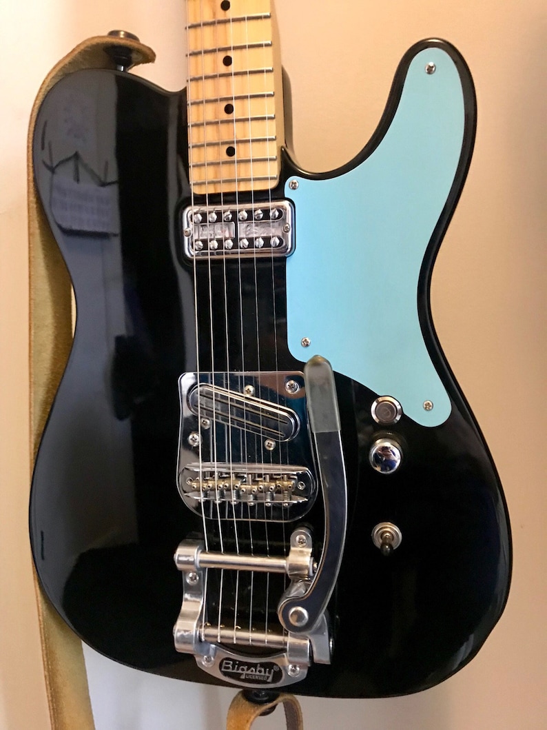 Puede incluir: Guitarra el&eacute;ctrica negra con una golpeador azul claro y un vibrato Bigsby.