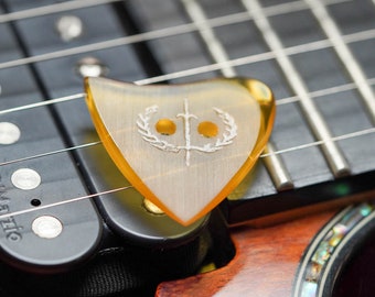 May include: A close-up of a yellow guitar pick with a white design of a sword and laurel wreath. The pick is resting on the strings of a guitar.