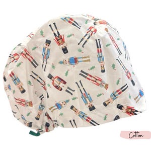 May include: White cotton cap featuring a repeating pattern of colourful nutcracker figures and green holly sprigs. The cap has a rounded crown and a gathered back. The word "Cotton" is printed on the bottom right.