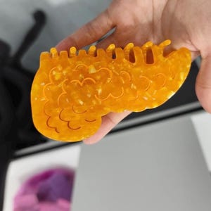 May include: A decorative hair clip in a warm amber color, held in a hand. The clip has a unique, organic shape with a series of curved cutouts and a textured surface. The clip is made of a translucent material with flecks of lighter color.